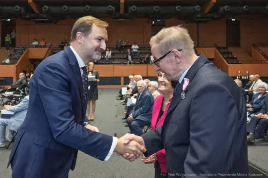 Uścisk dłoni dwóch mężczyzn w formalnym otoczeniu, w tle starsze osoby siedzące na widowni.