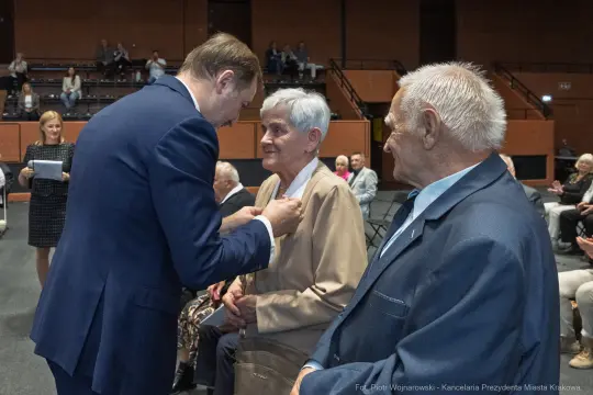 Uroczystość wręczenia odznaczenia starszej kobiecie przez mężczyznę, w tle siedzący goście.