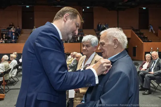 Uroczystość wręczenia odznaczeń, mężczyzna przypina medal starszemu mężczyźnie.
