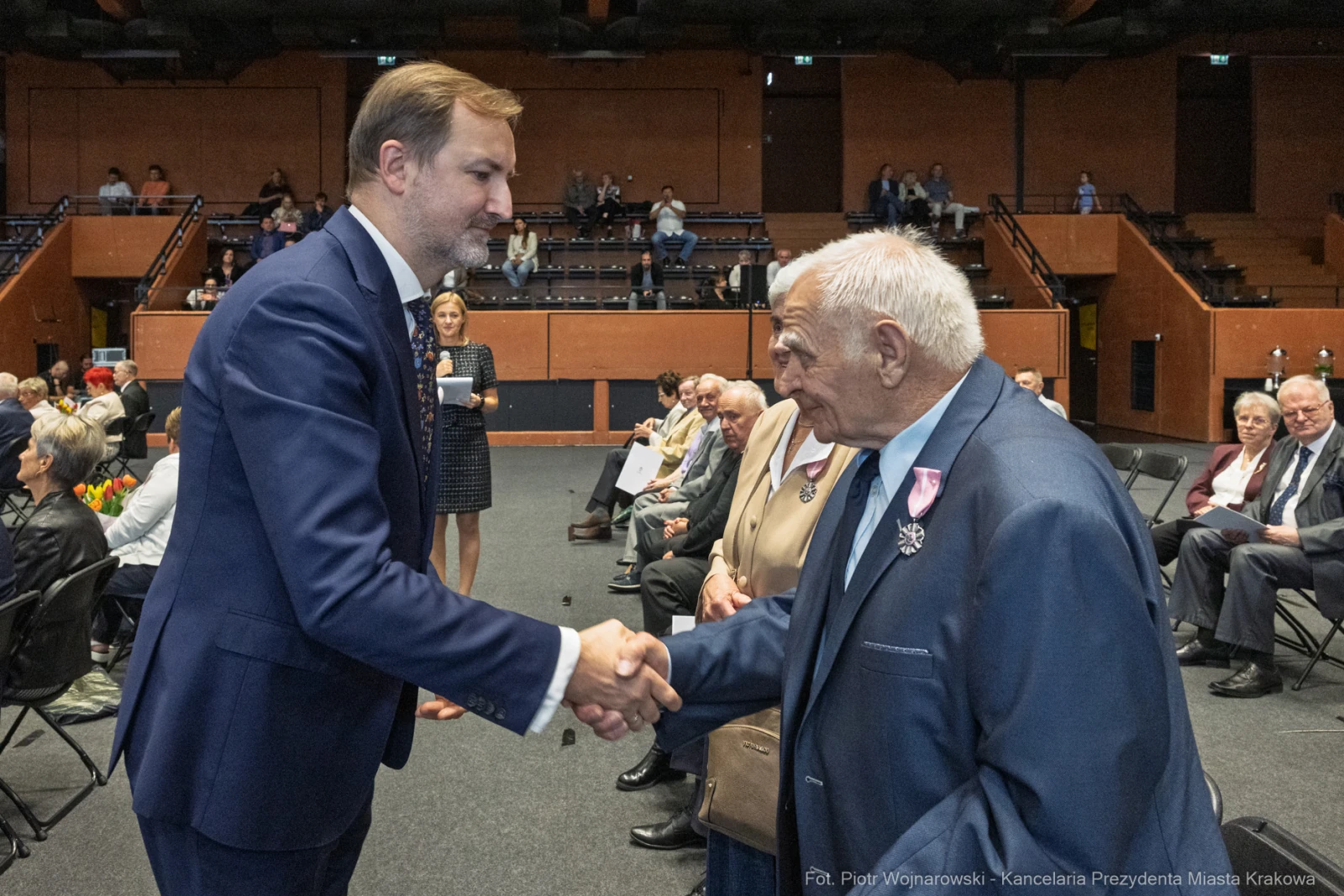 Uścisk dłoni między dwoma mężczyznami podczas uroczystości w sali. W tle siedzący goście.  Autor: Piotr Wojnarowski