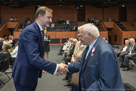 Uścisk dłoni między dwoma mężczyznami podczas uroczystości w sali. W tle siedzący goście.