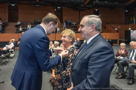 Wręczenie odznaczenia przez mężczyznę kobiecie w towarzystwie drugiego mężczyzny na uroczystości.