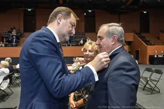 Wręczenie odznaczenia mężczyźnie przez innego mężczyznę, w tle kobieta i publiczność.