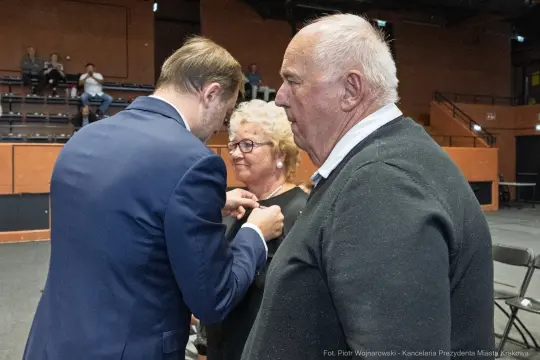 Wręczenie odznaczenia mężczyźnie przez osobę w garniturze oraz uśmiechnięta kobieta obok.