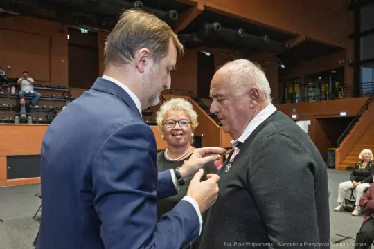 Uroczystość wręczenia odznaczenia mężczyźnie przez mężczyznę, z kobietą w tle.