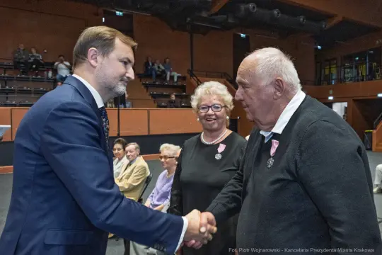 Uścisk dłoni mężczyzny z odznaczeniem, w tle starsze osoby siedzące na widowni.