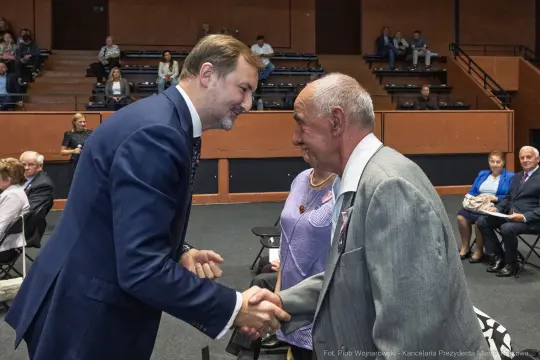 Uścisk dłoni dwóch mężczyzn podczas wydarzenia w sali. W tle widoczni uczestnicy.
