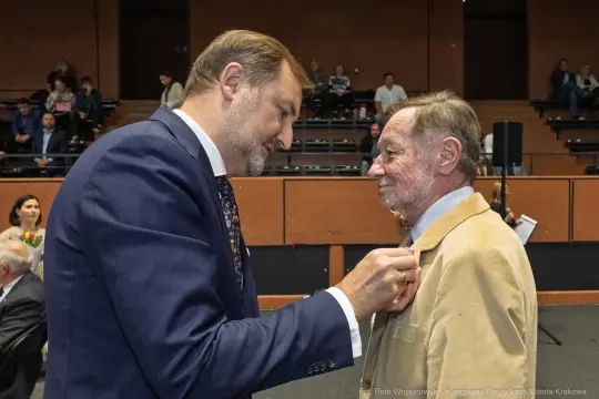 Wręczenie odznaczenia mężczyźnie przez innego mężczyznę w sali z widownią.
