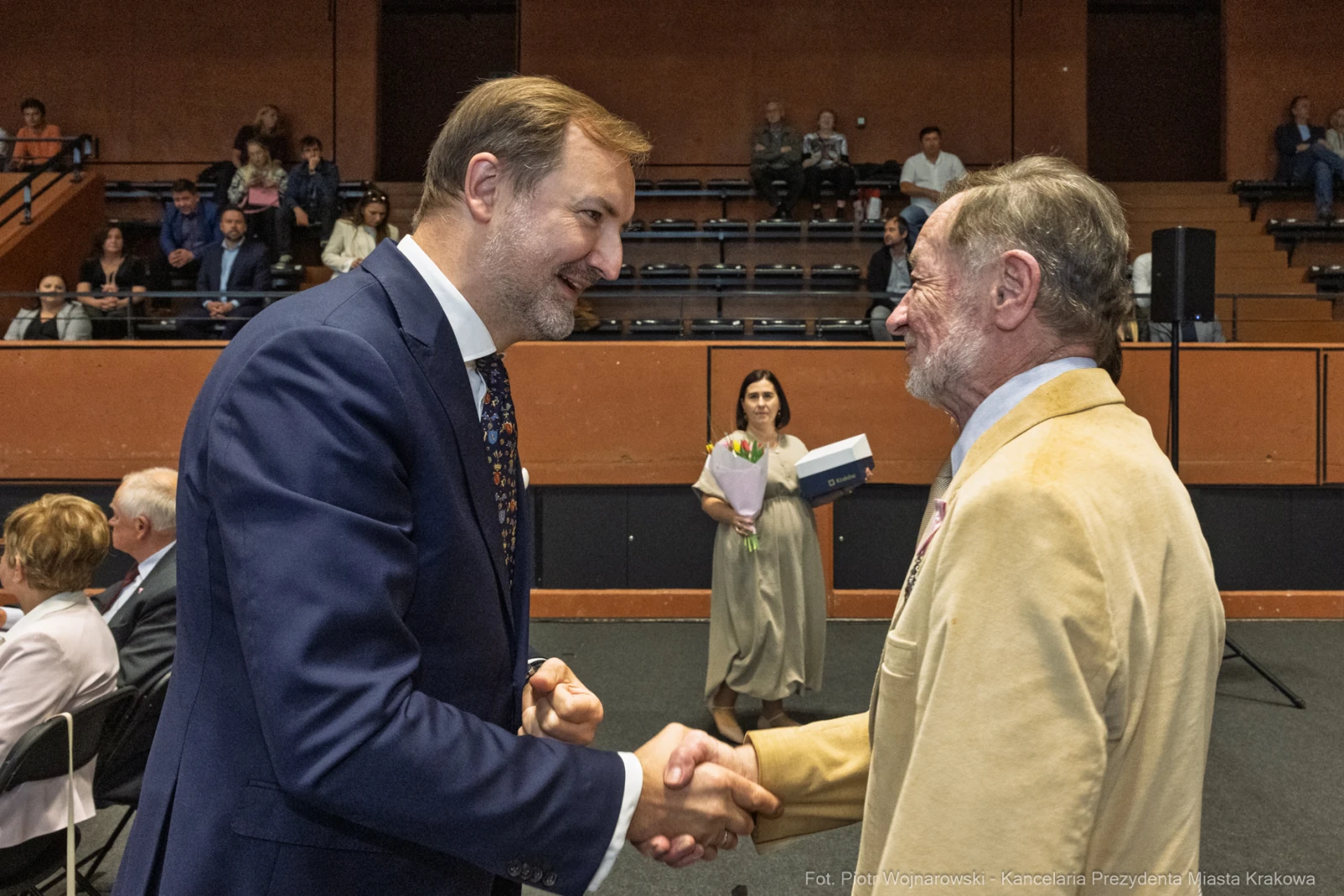 Dwaj mężczyźni uścisnęli sobie dłonie podczas uroczystości w sali. W tle widownia.  Autor: Piotr Wojnarowski