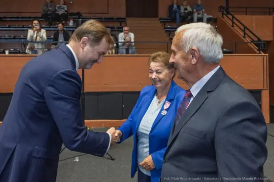 Uścisk dłoni między mężczyzną a kobietą w niebieskim garniturze, obok starszy mężczyzna.