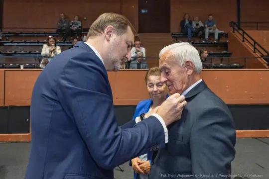 Wręczenie odznaczenia mężczyźnie w garniturze, obok kobieta i inny mężczyzna.