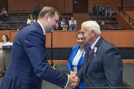 Uścisk dłoni dwóch mężczyzn, w tle kobieta i widownia. Wszyscy w formalnych strojach.