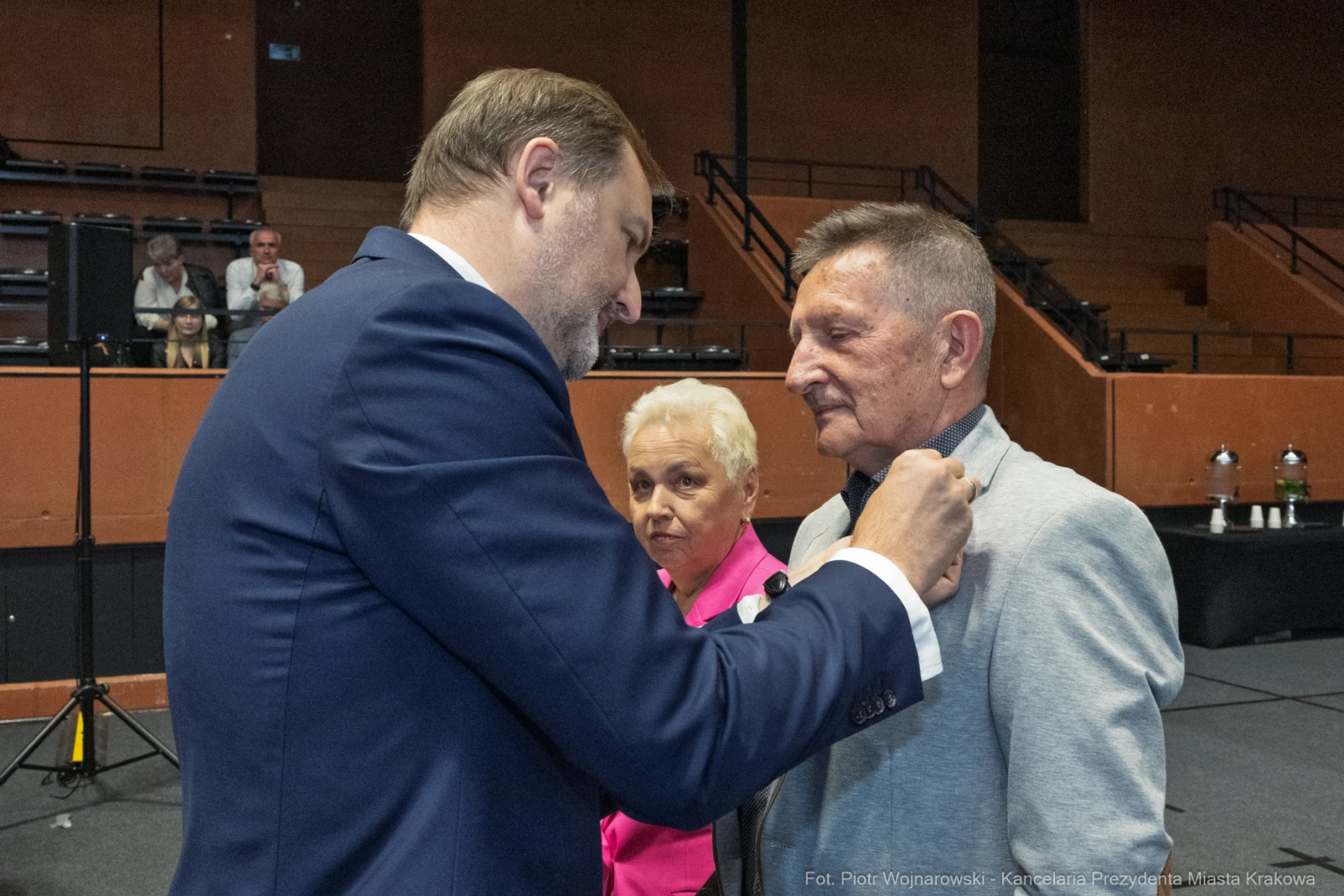 Wręczenie odznaczenia mężczyźnie przez elegancko ubranego mężczyznę, w tle kobieta.  Autor: Piotr Wojnarowski