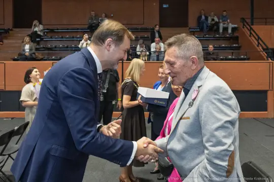 Uścisk dłoni dwóch mężczyzn podczas uroczystości w sali. W tle inne osoby w formalnych strojach.