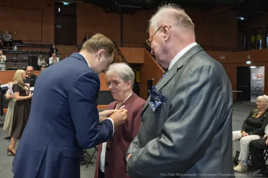 Wręczenie odznaczenia przez mężczyznę w garniturze kobiecie w okularach, obok stoi starszy mężczyzna.