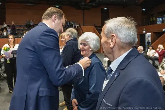Uroczystość wręczenia odznaczeń, osoby w eleganckich strojach, uśmiechy i gratulacje.