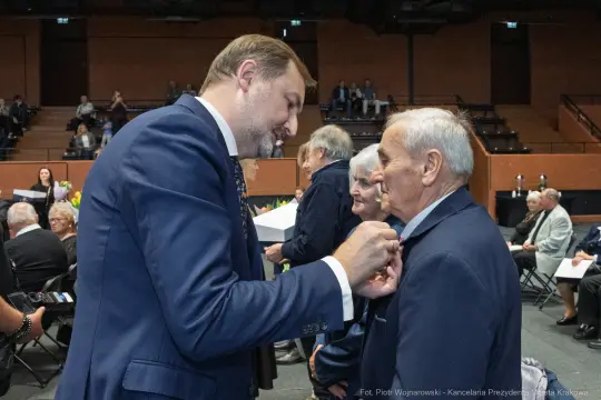 Mężczyzna przypina odznaczenie starszemu mężczyźnie w towarzystwie innych osób.