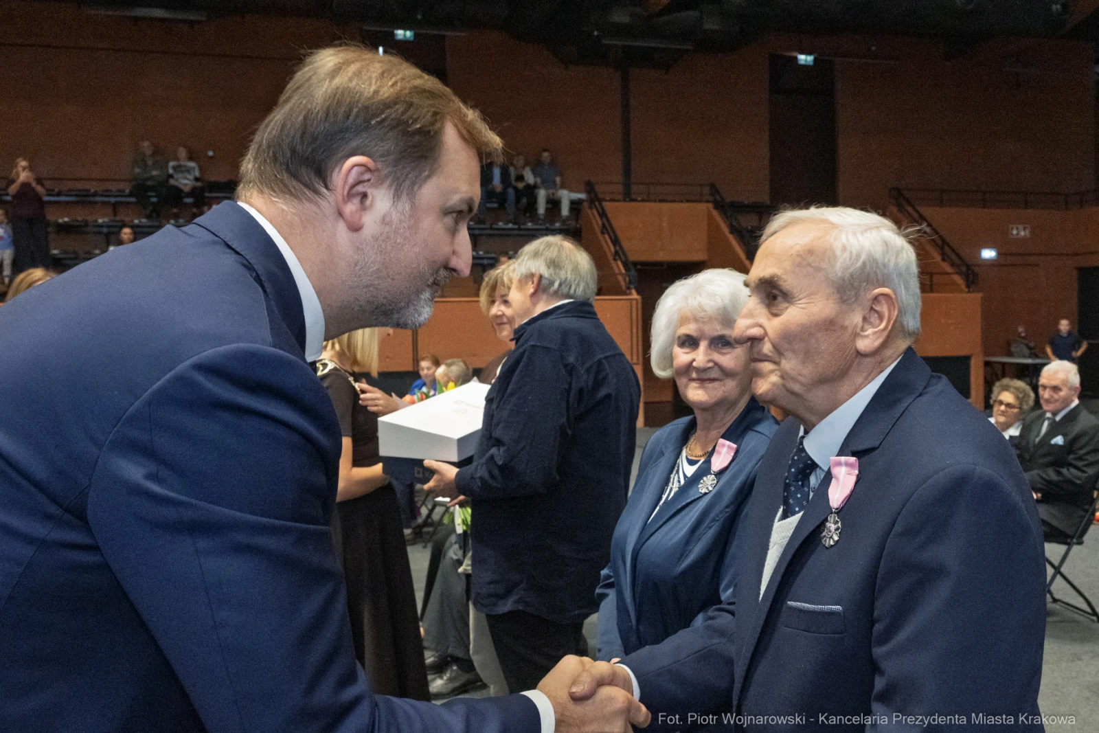 Uroczystość wręczenia odznaczeń, mężczyzna w garniturze podaje rękę starszemu mężczyźnie.  Autor: Piotr Wojnarowski