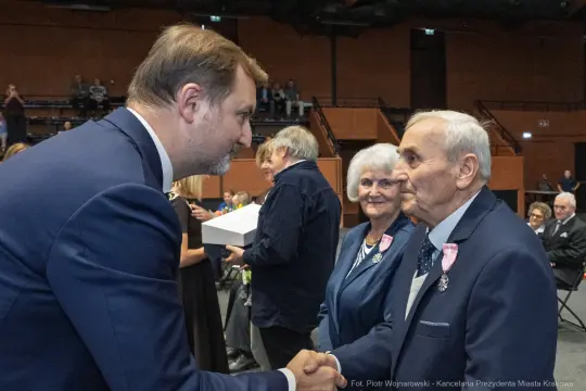 Uroczystość wręczenia odznaczeń, mężczyzna w garniturze podaje rękę starszemu mężczyźnie.