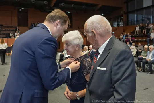 Wręczenie odznaczenia starszej kobiecie przez mężczyznę w garniturze, obok stoi starszy mężczyzna.