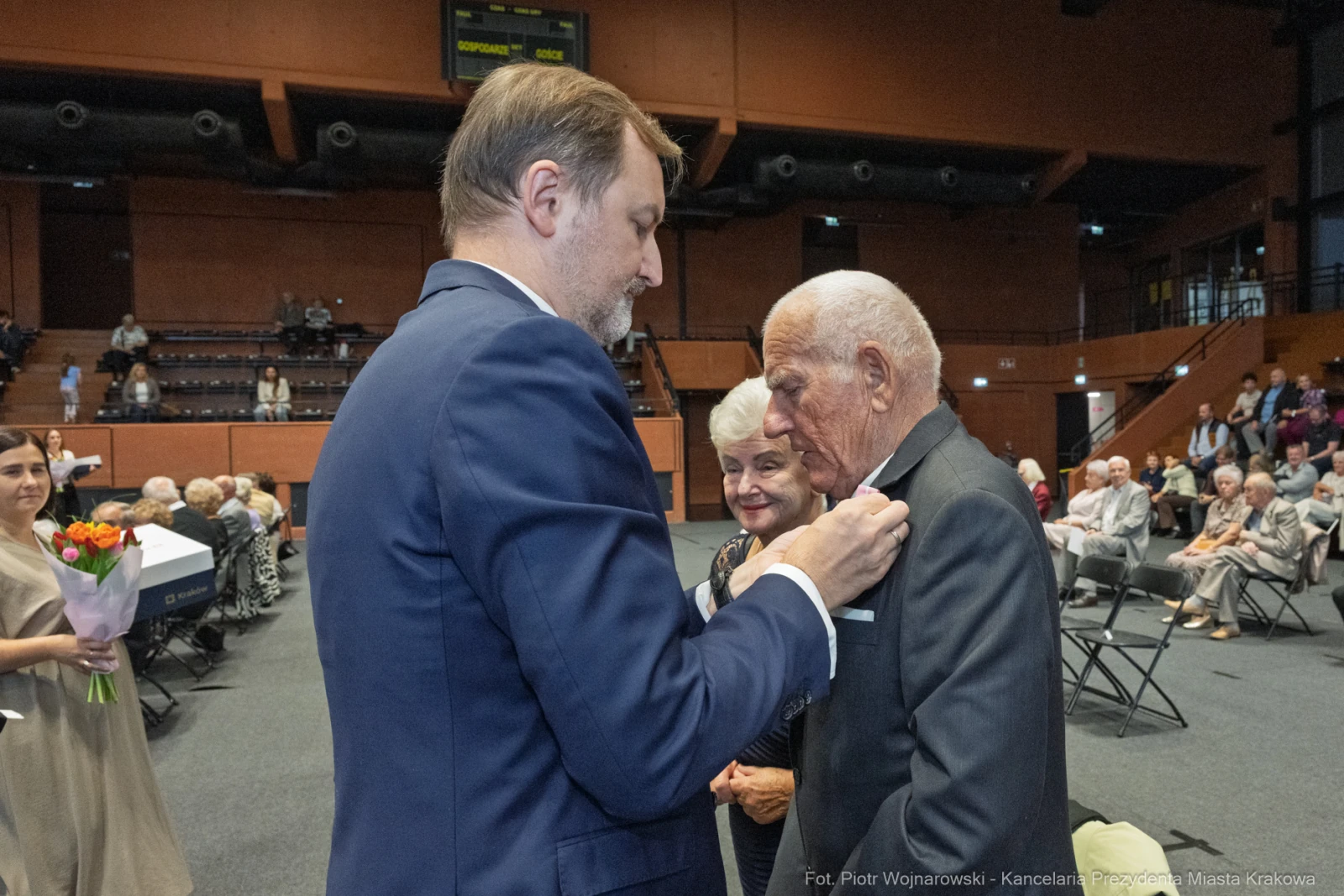 Uroczystość wręczenia odznaczeń, starszy mężczyzna w garniturze, uśmiechnięte osoby w tle.  Autor: Piotr Wojnarowski