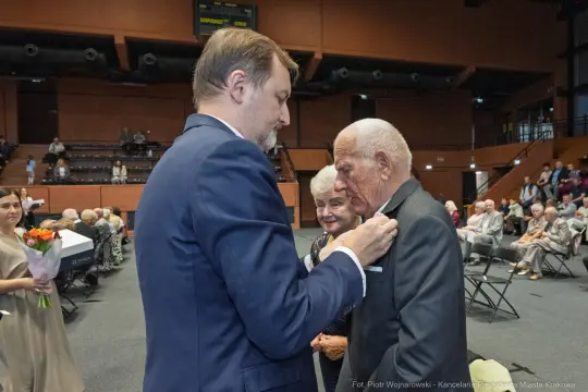 Uroczystość wręczenia odznaczeń, starszy mężczyzna w garniturze, uśmiechnięte osoby w tle.