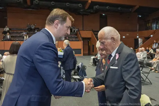 Uścisk dłoni dwóch mężczyzn podczas uroczystości, w tle siedzący goście.
