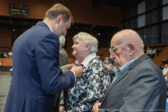 Wręczenie odznaczenia przez mężczyznę starszej kobiecie, obok siedzi starszy mężczyzna.