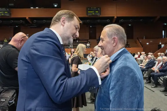 Mężczyzna przypina odznaczenie do marynarki innego mężczyzny podczas uroczystości.