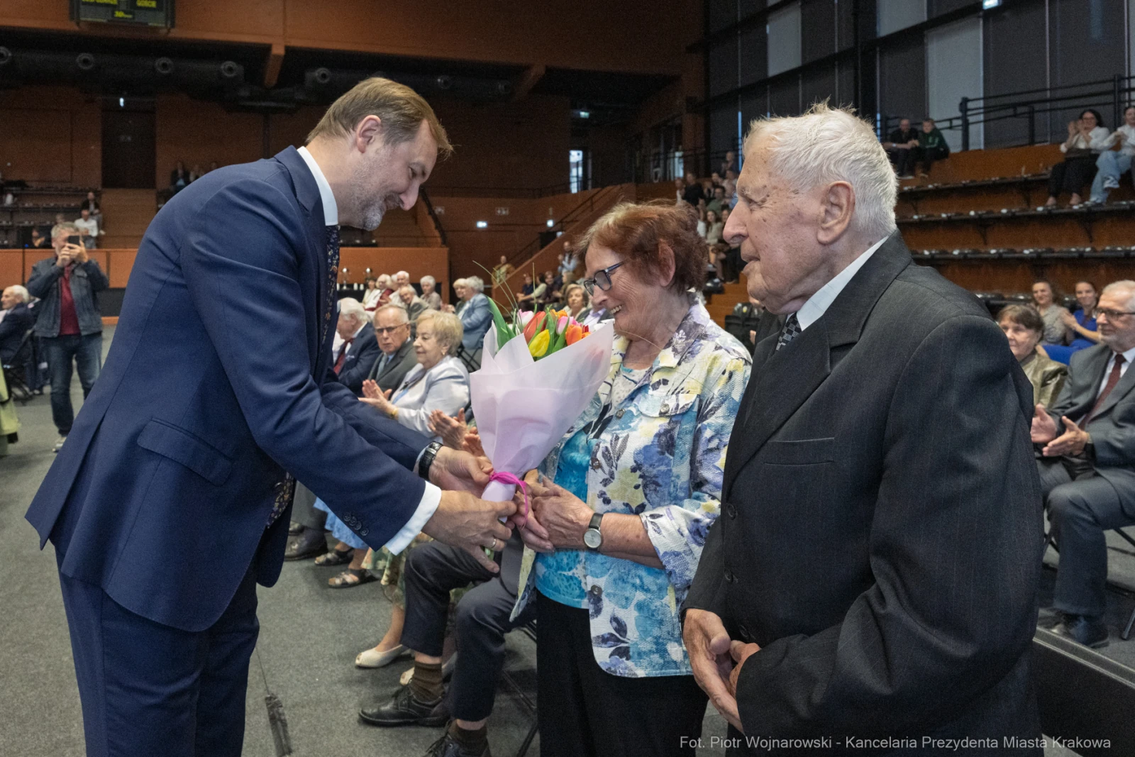 Mężczyzna wręcza bukiet kwiatów starszej kobiecie, obok stoi starszy mężczyzna. W tle widownia.  Autor: Piotr Wojnarowski