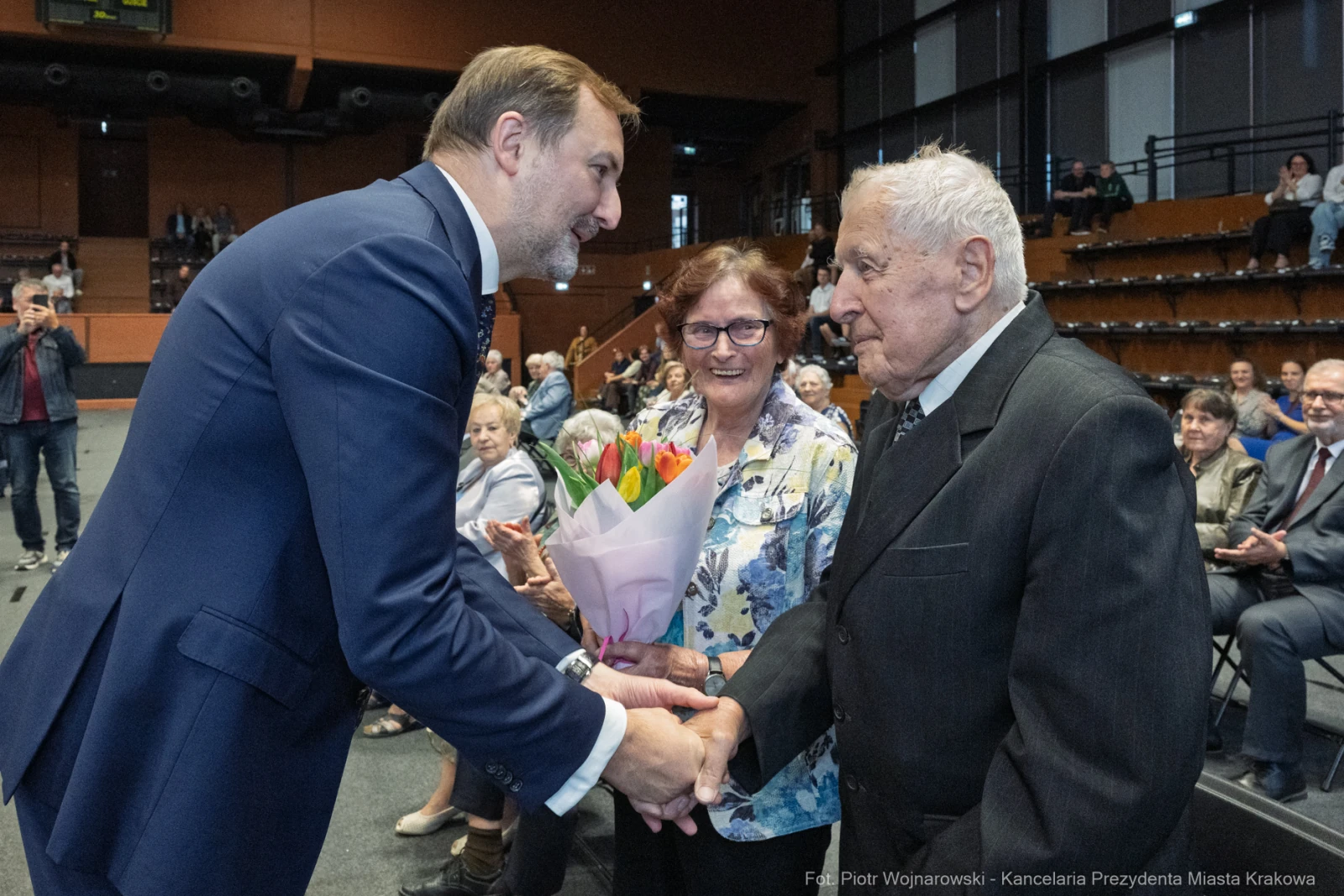 Mężczyzna wręcza bukiet kwiatów starszej parze w sali pełnej ludzi.  Autor: Piotr Wojnarowski