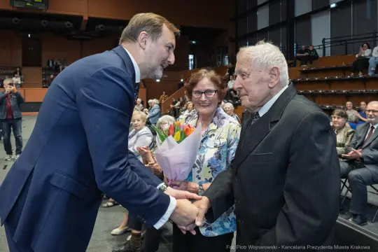 Mężczyzna wręcza bukiet kwiatów starszej parze w sali pełnej ludzi.