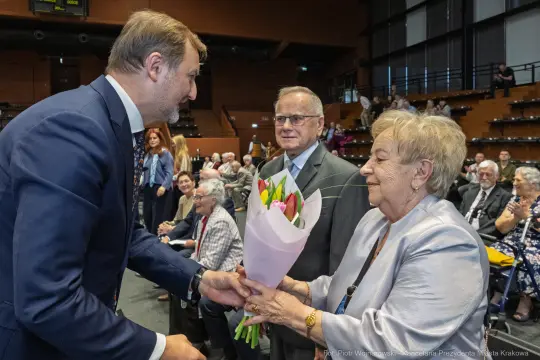 Mężczyzna wręcza bukiet kwiatów starszej kobiecie w towarzystwie innych osób.