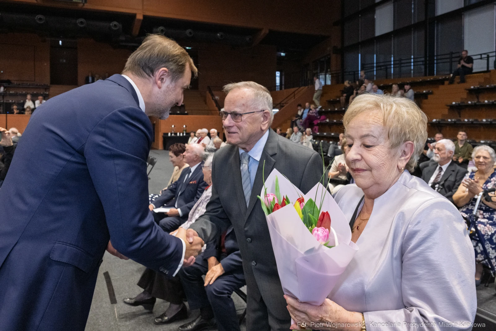 Uścisk dłoni, mężczyzna z bukietem kwiatów, starsza para w eleganckich strojach.  Autor: Piotr Wojnarowski