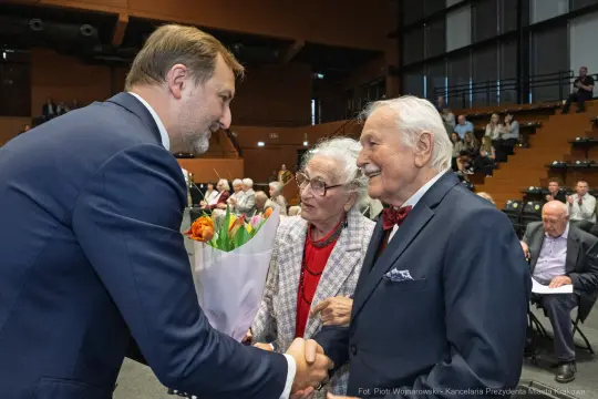 Uroczystość wręczenia kwiatów starszej parze przez mężczyznę w garniturze.
