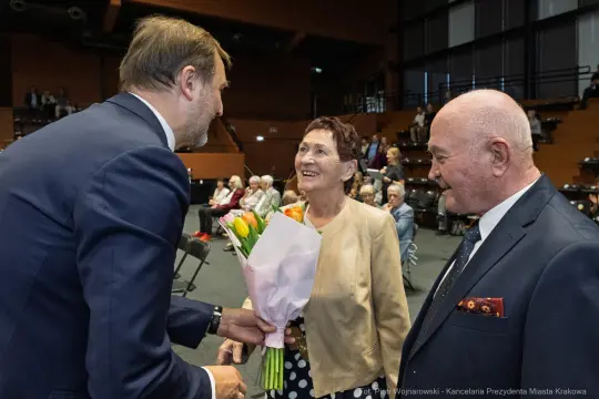 Mężczyzna wręcza bukiet kwiatów uśmiechającej się kobiecie w towarzystwie starszego mężczyzny.