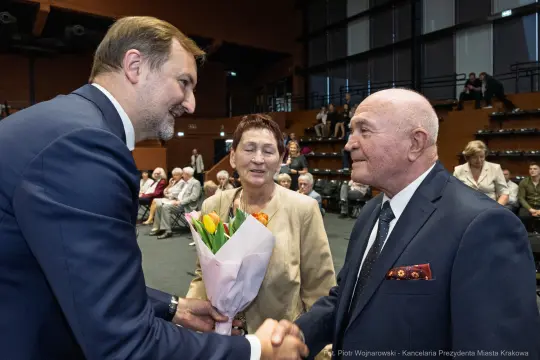 Mężczyzna w garniturze podaje rękę starszemu mężczyźnie, obok stoi kobieta z bukietem kwiatów.