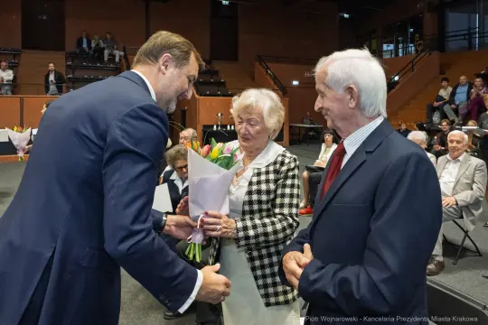 Uroczystość wręczenia kwiatów starszej parze przez mężczyznę w garniturze.