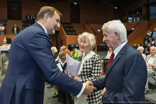 Wręczenie kwiatów przez mężczyznę starszej parze podczas uroczystości.