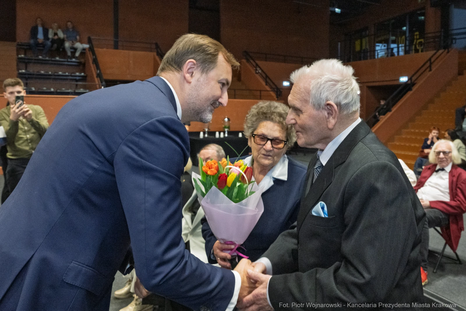 Uroczystość wręczenia kwiatów starszemu mężczyźnie przez młodszego mężczyznę.  Autor: Piotr Wojnarowski