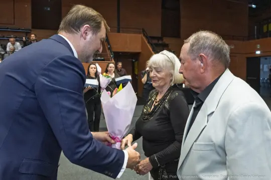 Mężczyzna wręcza bukiet kwiatów starszej kobiecie, obok stoi mężczyzna w jasnym garniturze.
