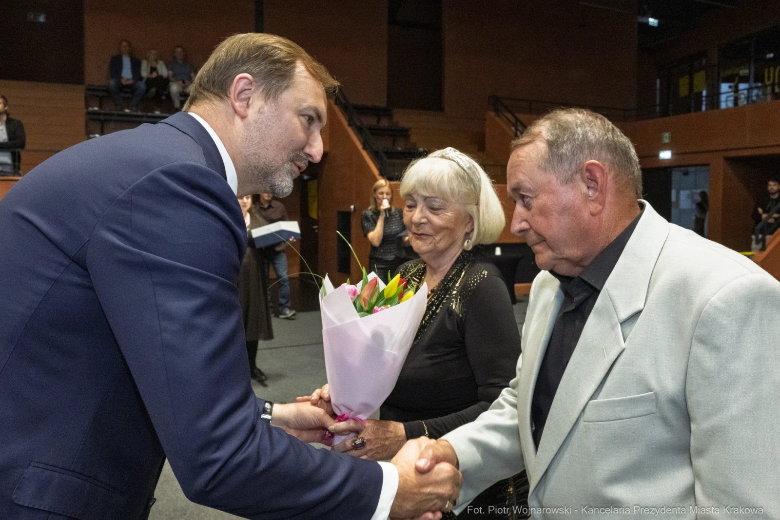 Uroczystość wręczenia kwiatów starszej parze przez mężczyznę w garniturze.  Autor: Piotr Wojnarowski