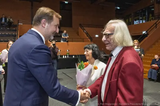 Spotkanie dwóch osób, jedna z bukietem kwiatów, w tle widownia.