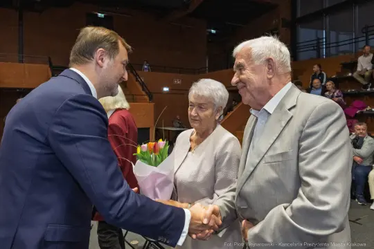 Uroczystość wręczenia kwiatów starszej parze przez mężczyznę w garniturze.