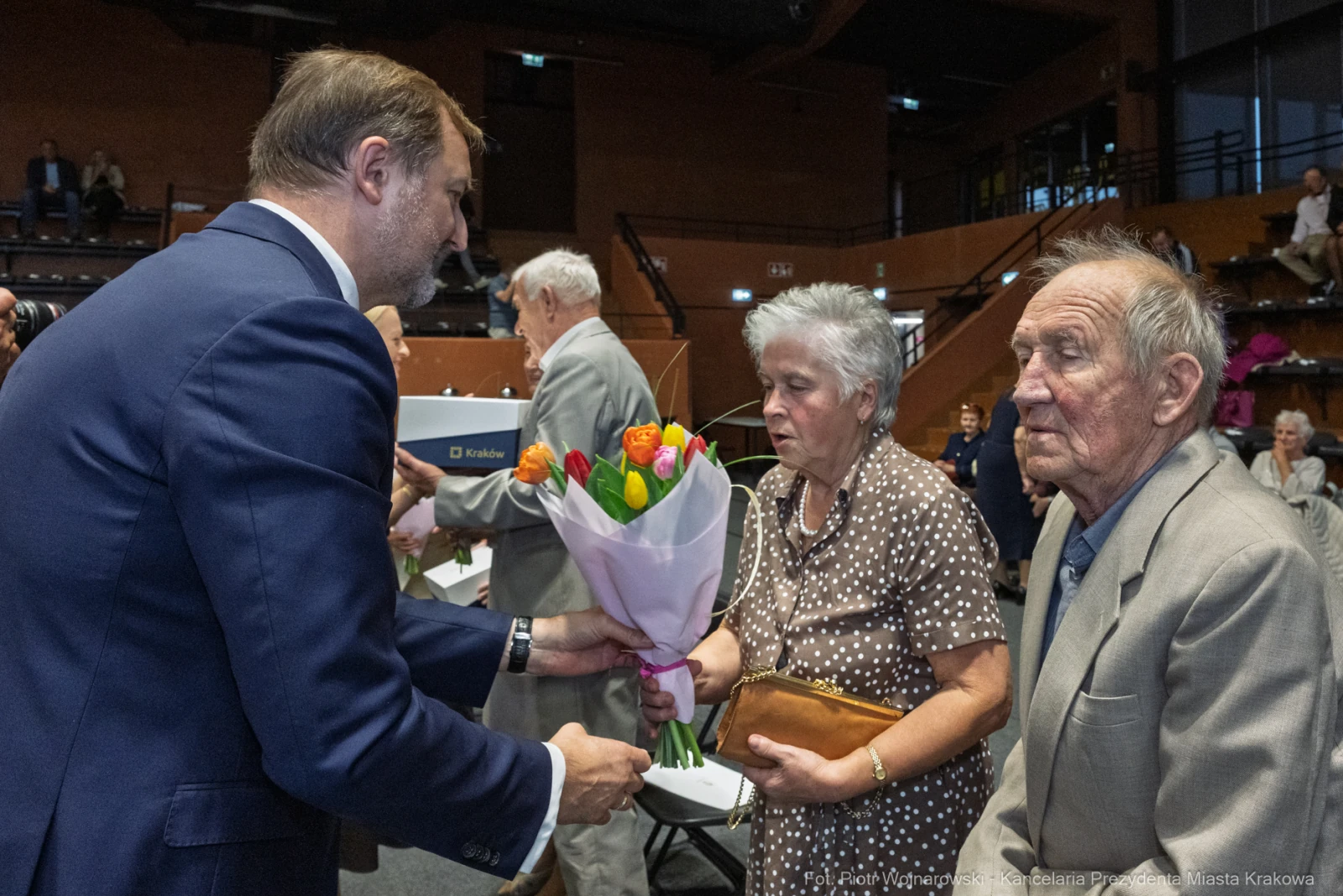 Wręczenie bukietu kwiatów starszej parze przez mężczyznę w garniturze.  Autor: Piotr Wojnarowski