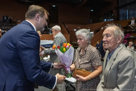 Wręczenie bukietu kwiatów starszej parze przez mężczyznę w garniturze.