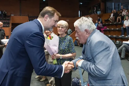 Uścisk dłoni między mężczyzną a starszym panem, w tle kobieta z bukietem kwiatów.