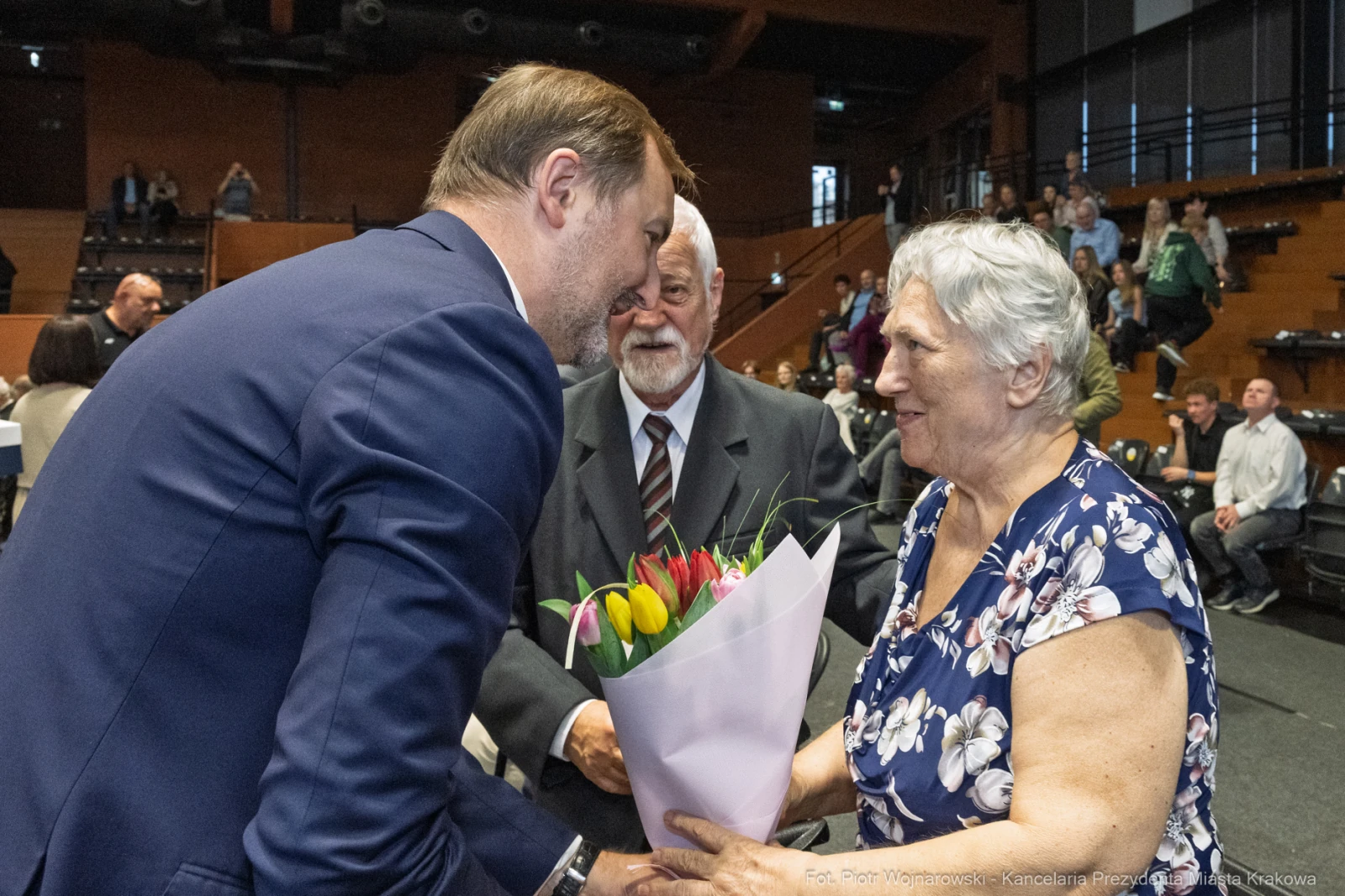 Mężczyzna wręcza bukiet kwiatów starszej kobiecie, obok stoi mężczyzna w garniturze.  Autor: Piotr Wojnarowski