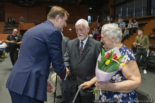 Mężczyzna w garniturze wita starsze małżeństwo, kobieta trzyma bukiet kwiatów.