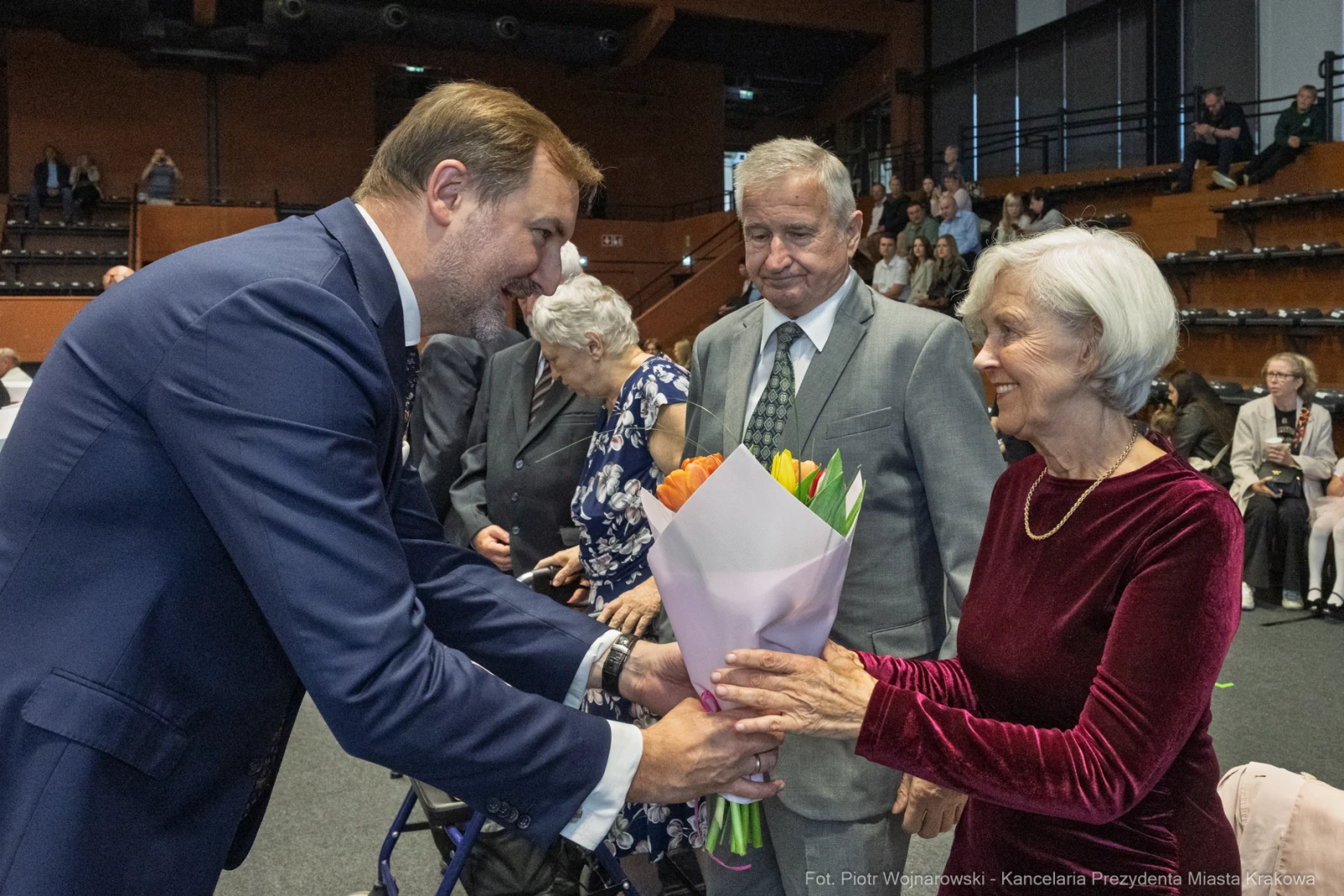 Mężczyzna wręcza bukiet kwiatów starszej kobiecie, w tle inni uczestnicy wydarzenia.  Autor: Piotr Wojnarowski